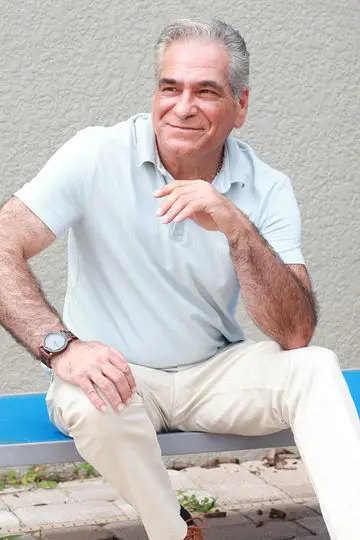 Phil Green seated outdoors on a blue bench in a light blue polo and khaki trousers, looking off-camera with a slight smile