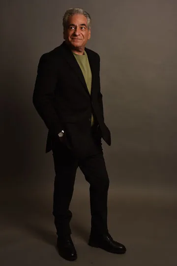 Full-body studio portrait of Phil Green in a black suit jacket and olive t-shirt, standing against a dark backdrop